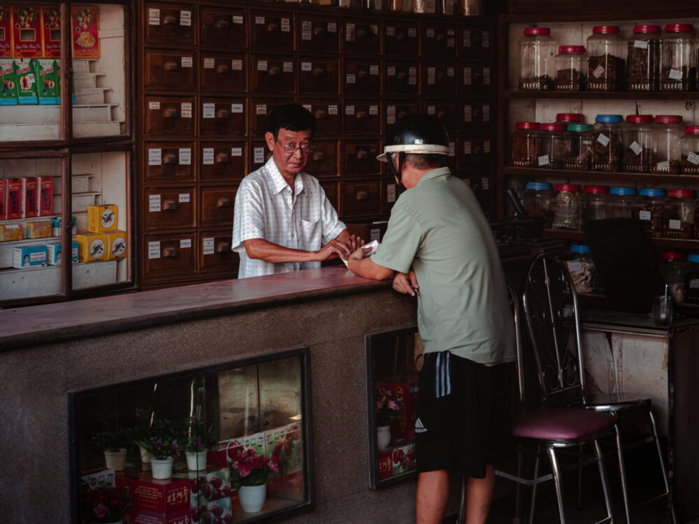 Two men speaking in a Vietnamese herbal medicine store with wooden drawers and glass jars.