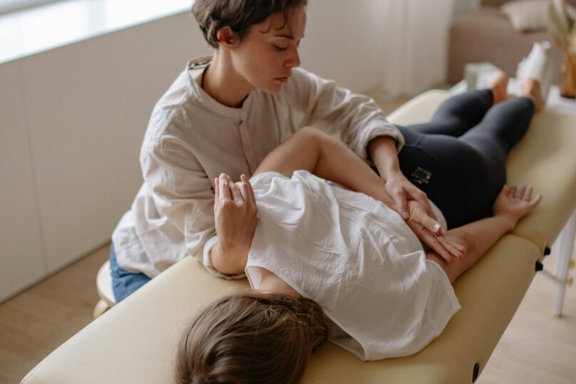A woman lying on a massage table receiving a chiropractic adjustment in a bright room.