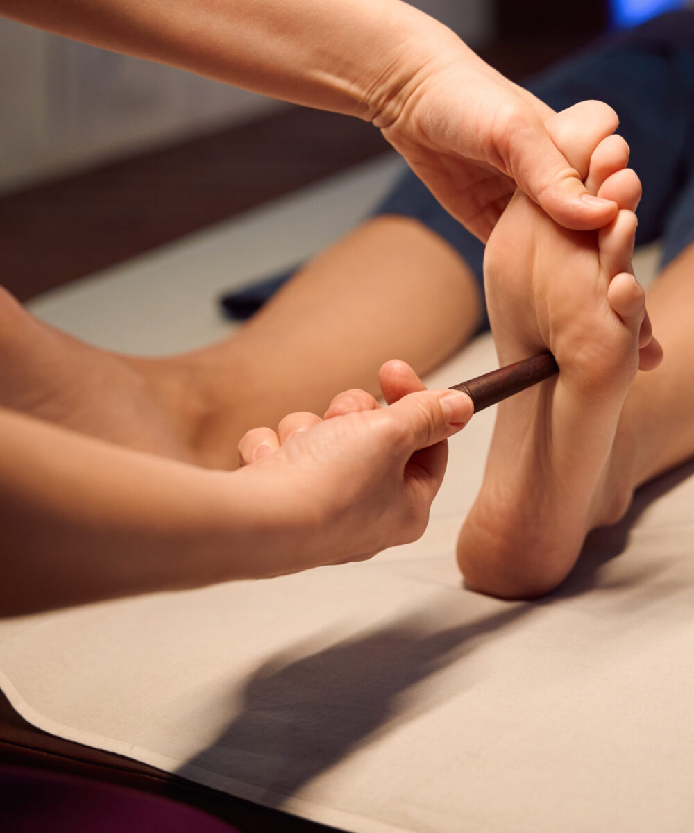 Cropped photo of masseuse applying pressure with wooden reflexology stick to reflex point on client foot arch